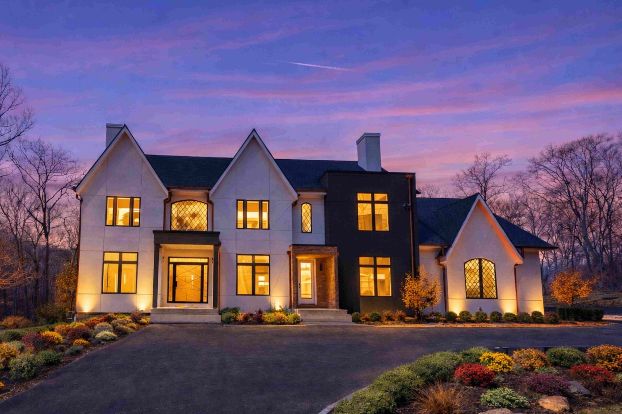 Dusk view of a modern tudor style custom home in Great Falls, VA featuring light-colored stucco and dark accents, symmetrical gables, warmly lit windows, a landscaped driveway and exterior, and mature trees surrounding the property.